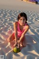 A woman in a pink bathing suit sitting on a beach with a coconut.